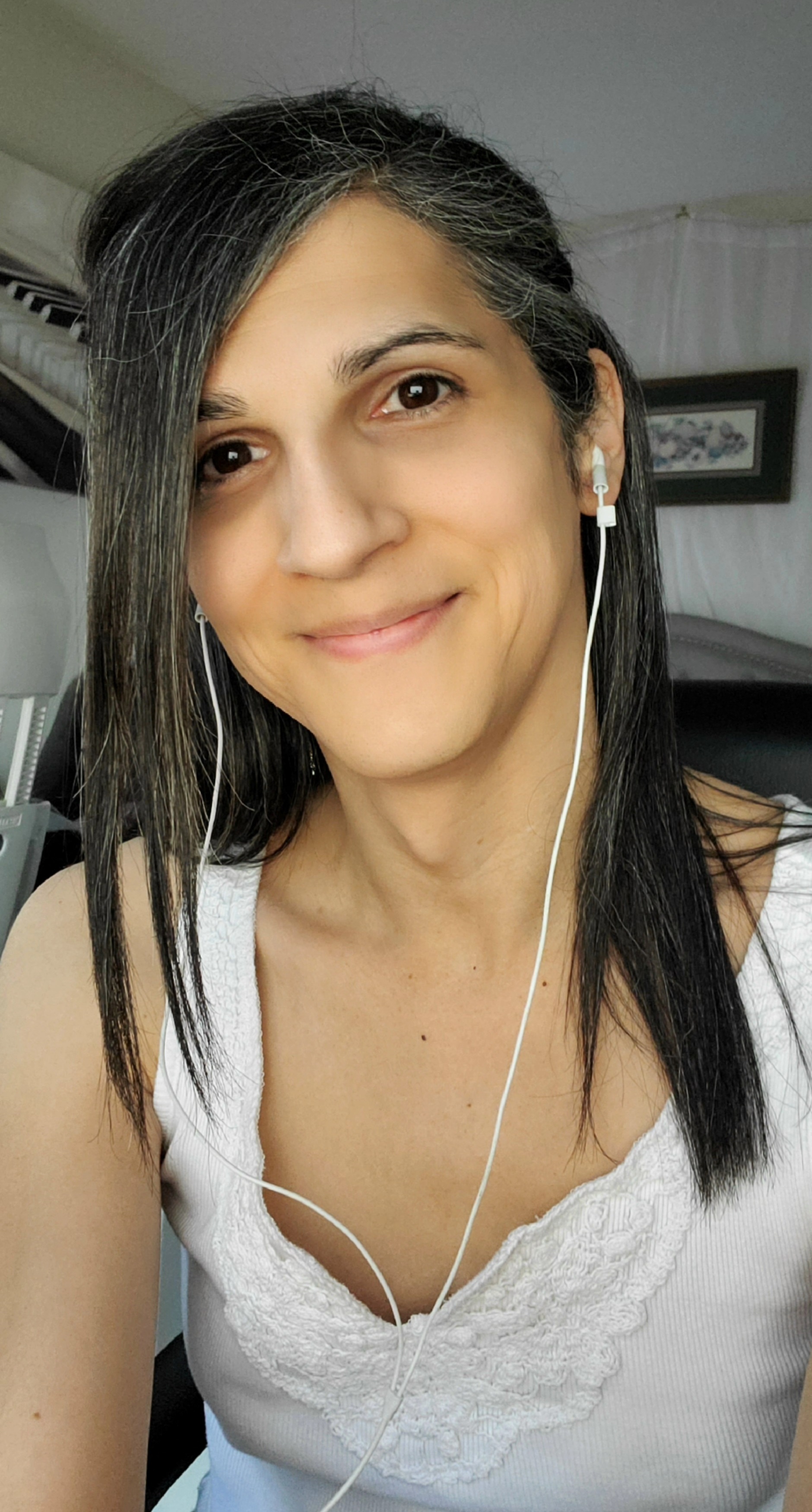 A picture of a tan, dark-haired, smiling woman in a white top. The woman is wearing ear buds. Two pictures are hanging on the walls behind her, visible over her shoulders. The picture to the left of the woman depicts a piano keyboard. The picture to the right of the woman is a brown-framed floral print, though the picture is far enough in the camera's depth of field to be blurry and difficult to discern.